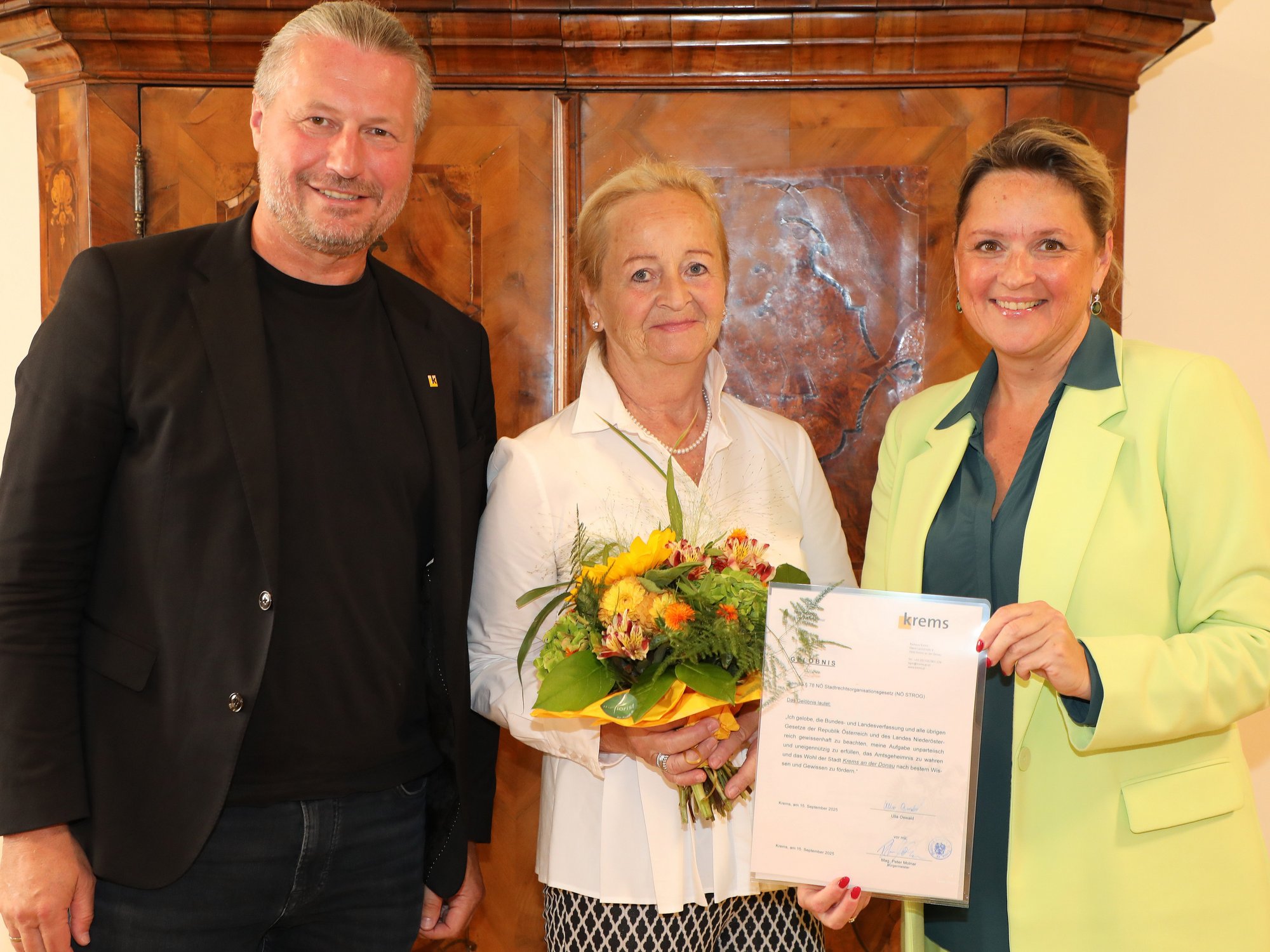 Drei Personen vor einen historischen Möbelstück, eine Frau hält einen Blumenstrauß, eine weitere eine Urkunde mit Logo der Stadt Krems