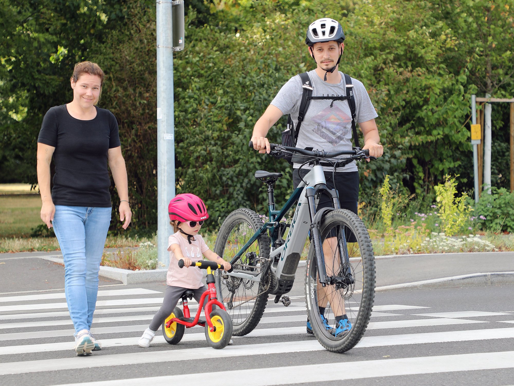 Mann mit E-Bike, Kind mit Laufrad und Frau auf einem Zebrastreifen