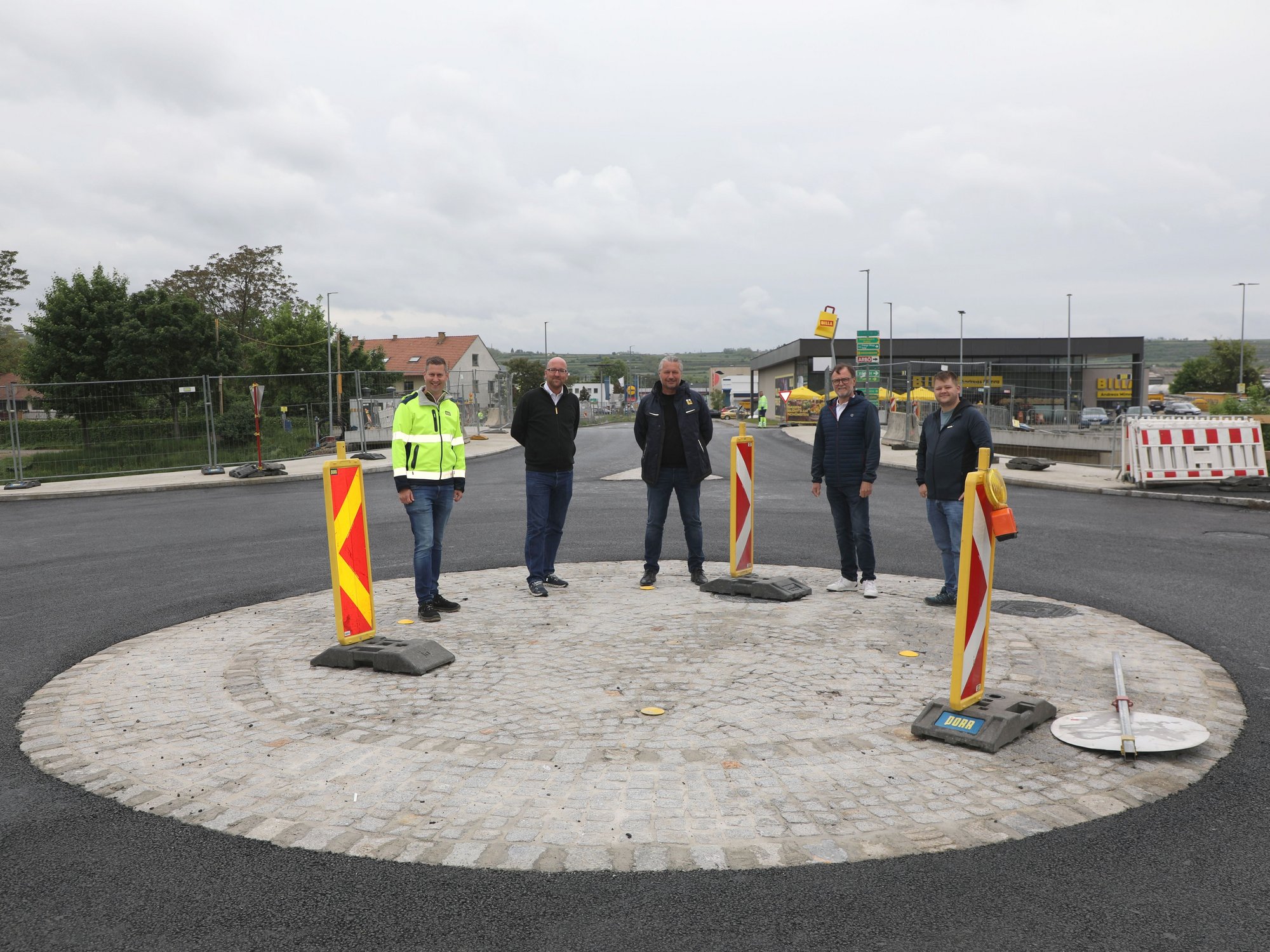Fünf Männer stehen in der Mitte eines Kreisverkehrs, im Hintergrund sind eine Brücke sowie ein Billa-Supermarkt zu sehen