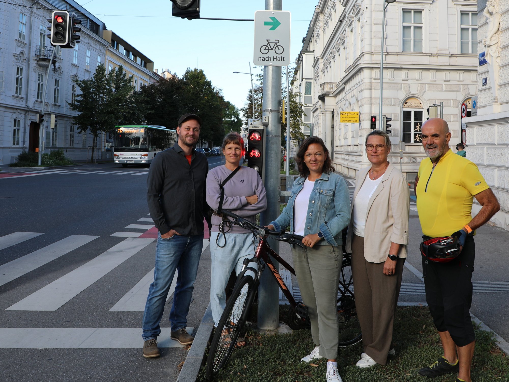 Fünf Personen stehen mit einem Fahrrad bei einem Verkehrsschild mit dem Hinweis „nach Halt“ an einer Kreuzung in städtischer Umgebung.