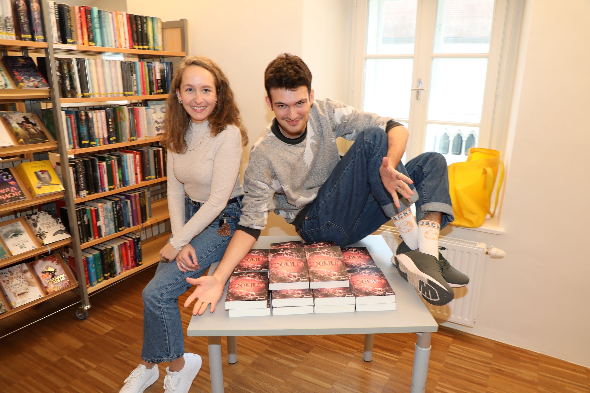 Autor Colin Hadler und Büchereileiterin Kathrin Raffetseder sitzend auf einem Büchertisch