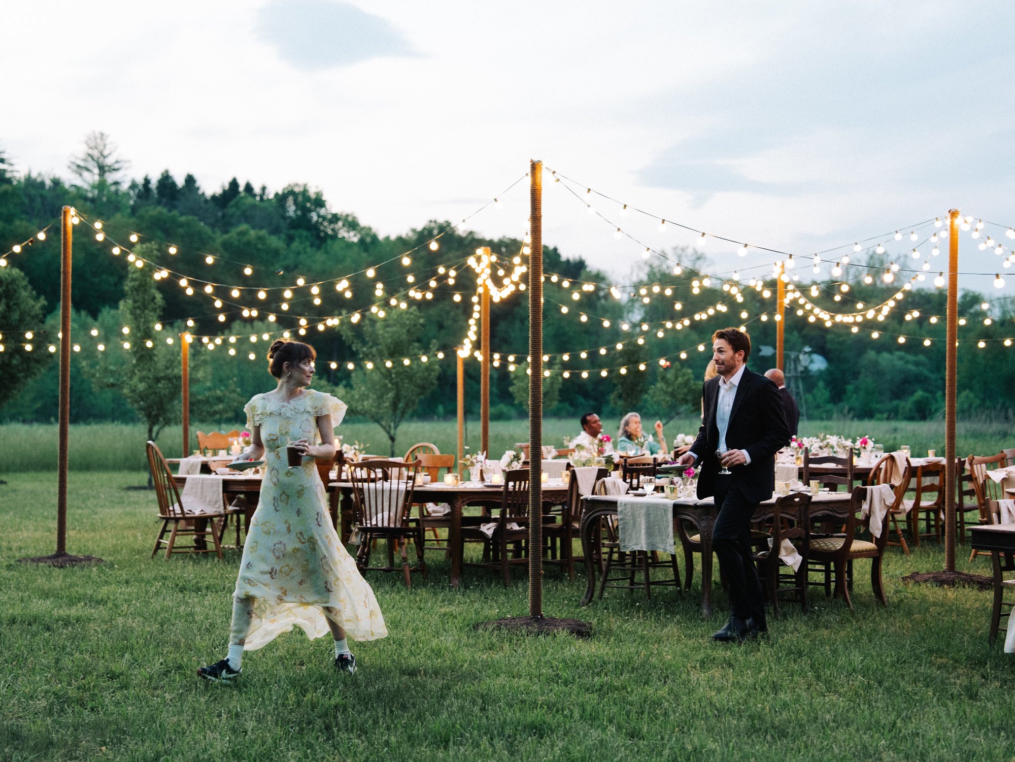 Filmszene: Feier im Freien mit Lichterketten, gedeckten Tischen und Gästen auf einer Wiese.