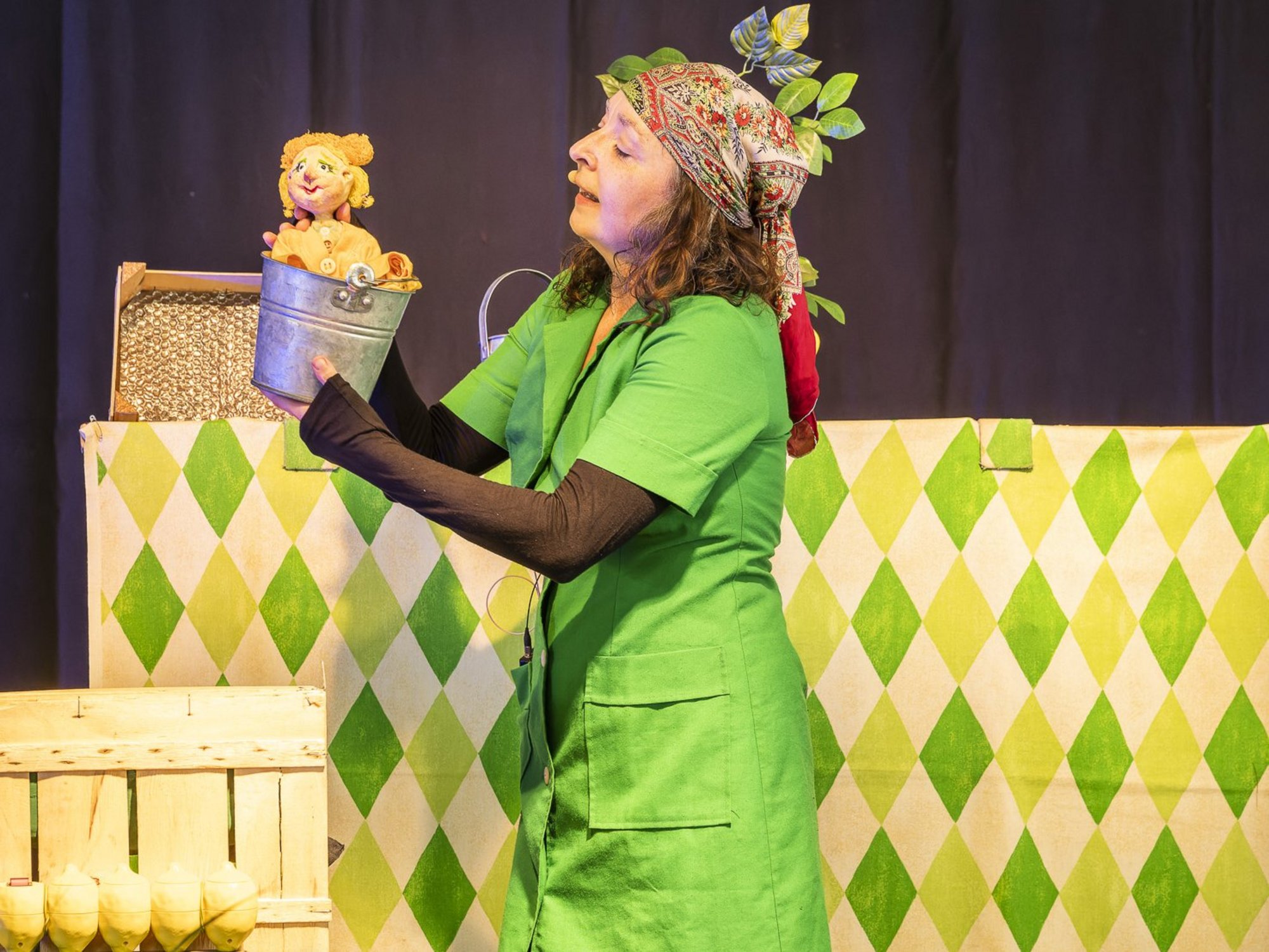 Frau mit grünem Kostüm und Kopftuch hält eine Handpuppe in einem Blecheimer auf einer Bühnenkulisse.