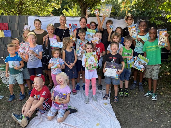Alt-Texte sagte:  Gruppe von Kindern und Erwachsenen mit Büchern und Urkunden bei einer Veranstaltung im Freien.